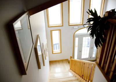 Bright wooden stairwell at Stay by the Sea B&B in Iqaluit, Nunavut, seen from the second floor looking down toward the main entrance with large windows and framed artwork along the wall.