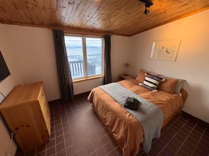 Room 5 at Stay by the Sea B&B in Iqaluit featuring a cozy queen bed, wooden dresser, and a scenic view of Frobisher Bay through the large window.