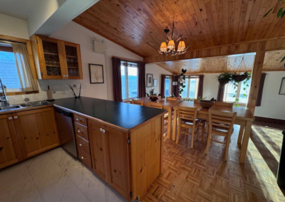 Open-concept common kitchen and dining area at Stay by the Sea B&B in Iqaluit, featuring wood cabinetry, a large dining table, hanging plants, and ocean views through bright windows.