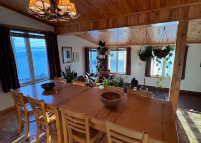 Bright dining room at Stay by the Sea B&B in Iqaluit, Nunavut, featuring a large wooden table, natural light, indoor plants, and panoramic views of Frobisher Bay through picture windows.
